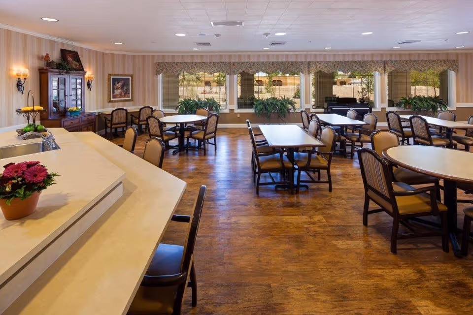 A spacious dining room with multiple tables and chairs arranged neatly on a wooden floor. The room features large windows with valances, allowing natural light to brighten the space. There is a counter with a sink and a potted plant in the foreground, and a wooden cabinet with decorative items against the wall.