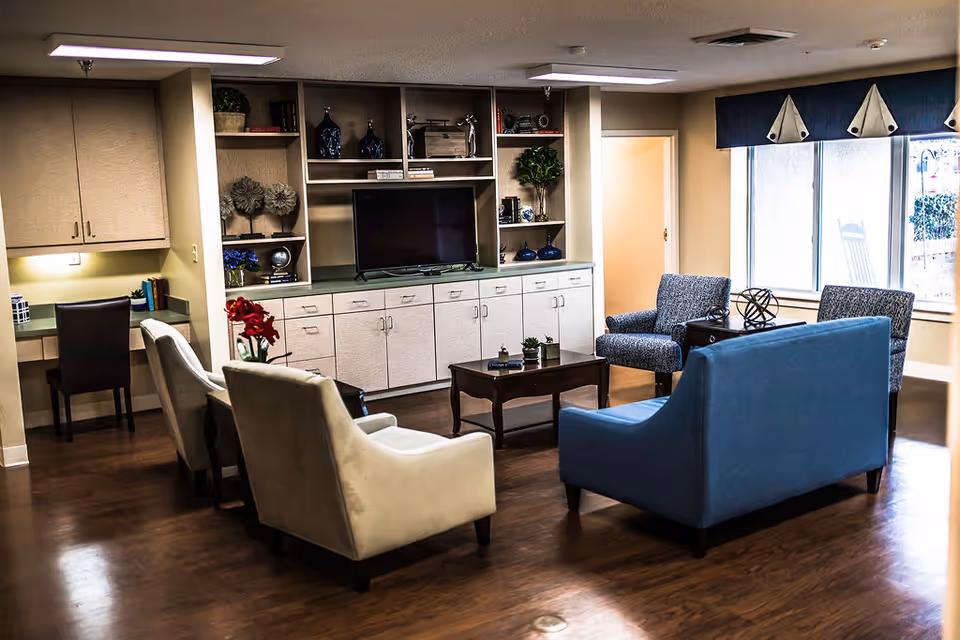 A cozy senior living facility common area with a blue loveseat, two beige armchairs, and two patterned armchairs arranged around a wooden coffee table. Behind the seating area is a built-in cabinet with shelves holding decorative items and a flat-screen TV. A desk with a chair is visible in the corner, and large windows with blue valances let in natural light.
