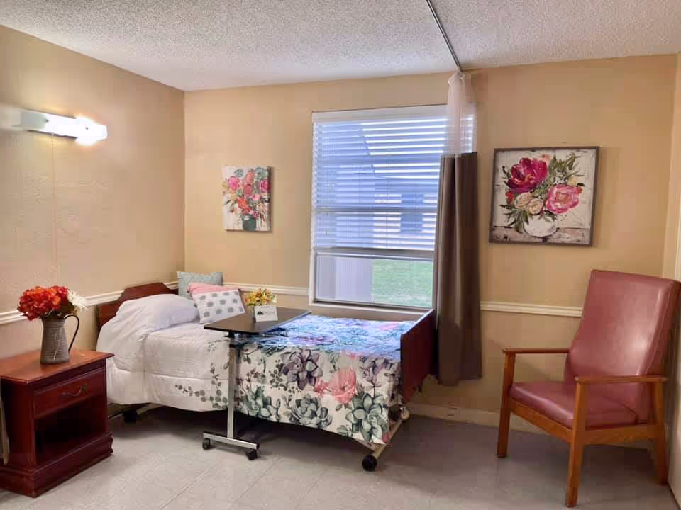 Small furnished bedroom with a single bed covered in floral bedding, an overbed table, nightstand with flowers, and a chair by a window.