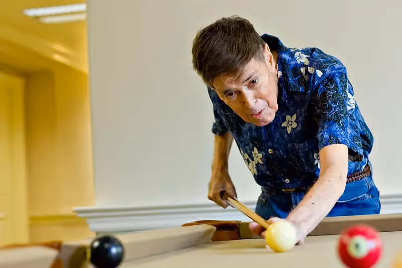 An elderly man wearing a blue floral shirt is playing pool indoors, aiming to strike the cue ball with a pool cue on a beige pool table.
