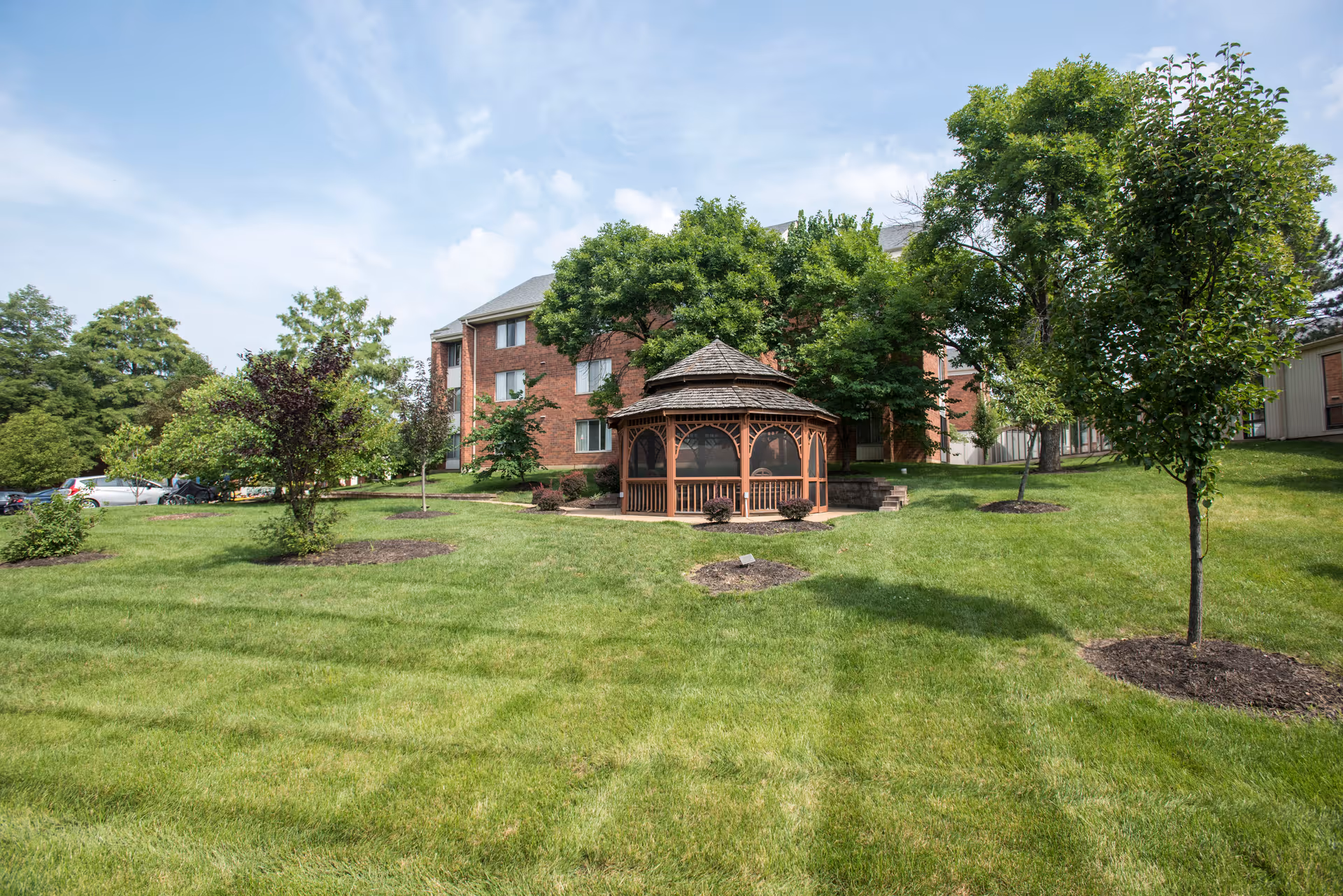 Brick senior living building with a wooden gazebo on a manicured lawn surrounded by trees and parked cars in the background.