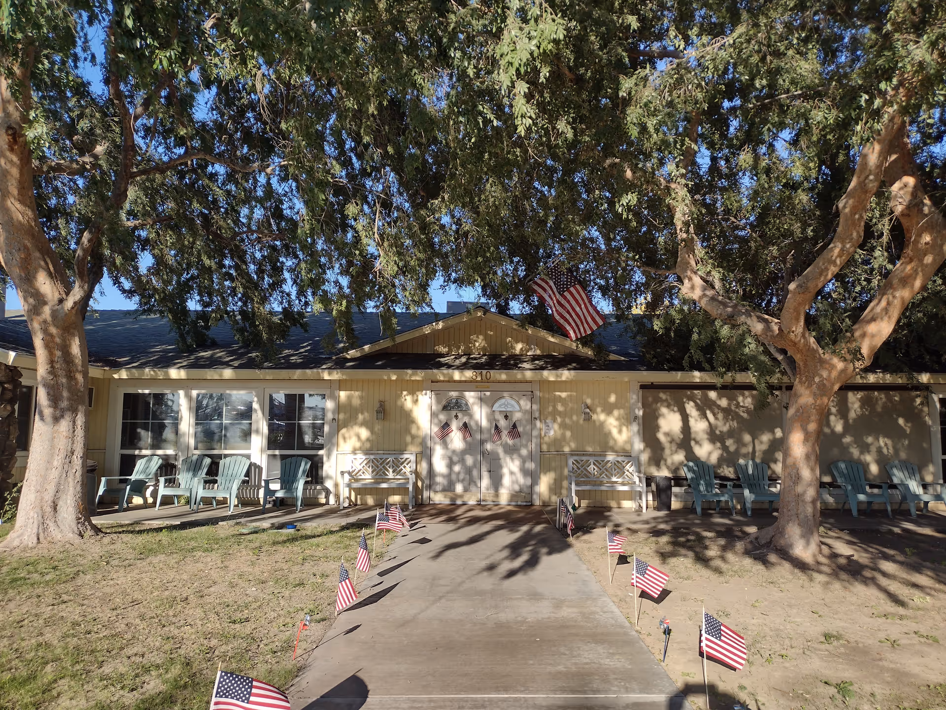 Front entrance of a single-story assisted living building with a walkway lined with small American flags, benches and chairs under large trees.