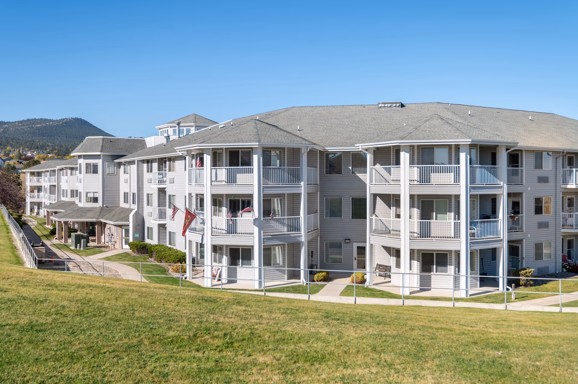 Exterior view of a multi-story senior living facility building with balconies, surrounded by a grassy area and a clear blue sky in the background.