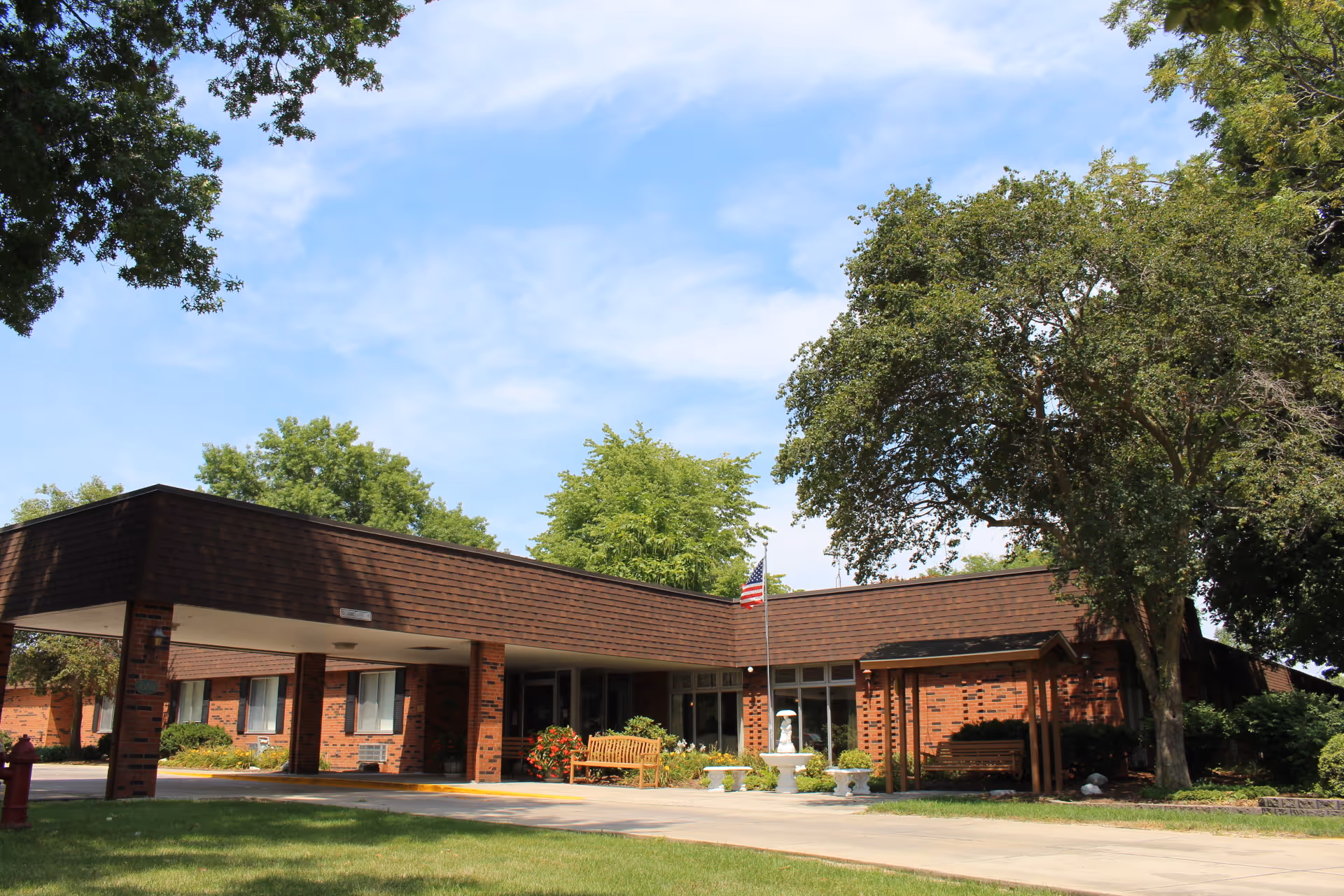 Exterior view of a single-story brick building with a covered entrance, surrounded by trees and greenery under a partly cloudy sky. There is an American flag on a flagpole near the entrance and benches placed outside.