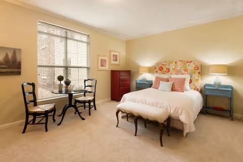 A cozy bedroom with a floral upholstered headboard, a white bedspread, and decorative pillows. There are two blue nightstands with lamps on either side of the bed, a red chest of drawers against the wall, and a small round table with two chairs near a window with blinds. The walls are painted beige and the floor is carpeted.