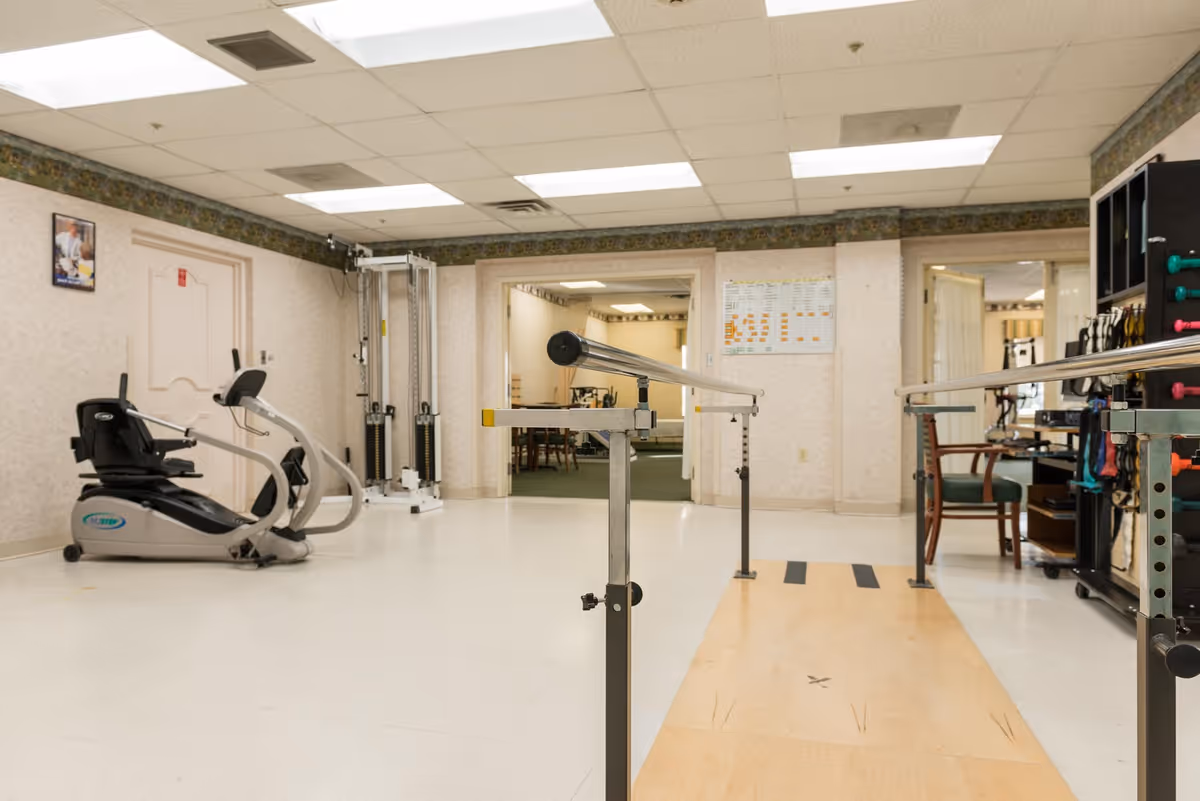 Rehabilitation gym room with parallel walking bars, an exercise bike, weight machines, and therapy equipment.
