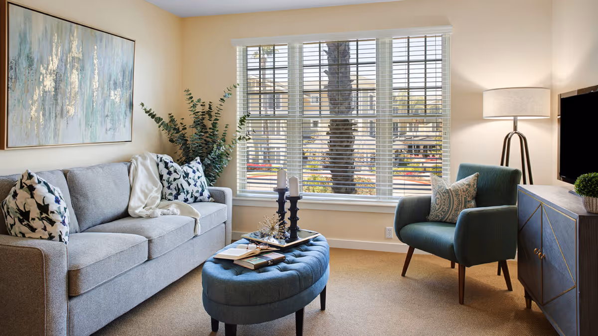 A cozy living room with a gray sofa adorned with patterned pillows and a white throw blanket. In front of the sofa is a blue tufted ottoman with books and decorative items on top. To the right, there is a teal armchair with a patterned pillow, a floor lamp, and a TV on a blue cabinet. Large windows with white blinds allow natural light to fill the room, showing a view of palm trees and buildings outside.