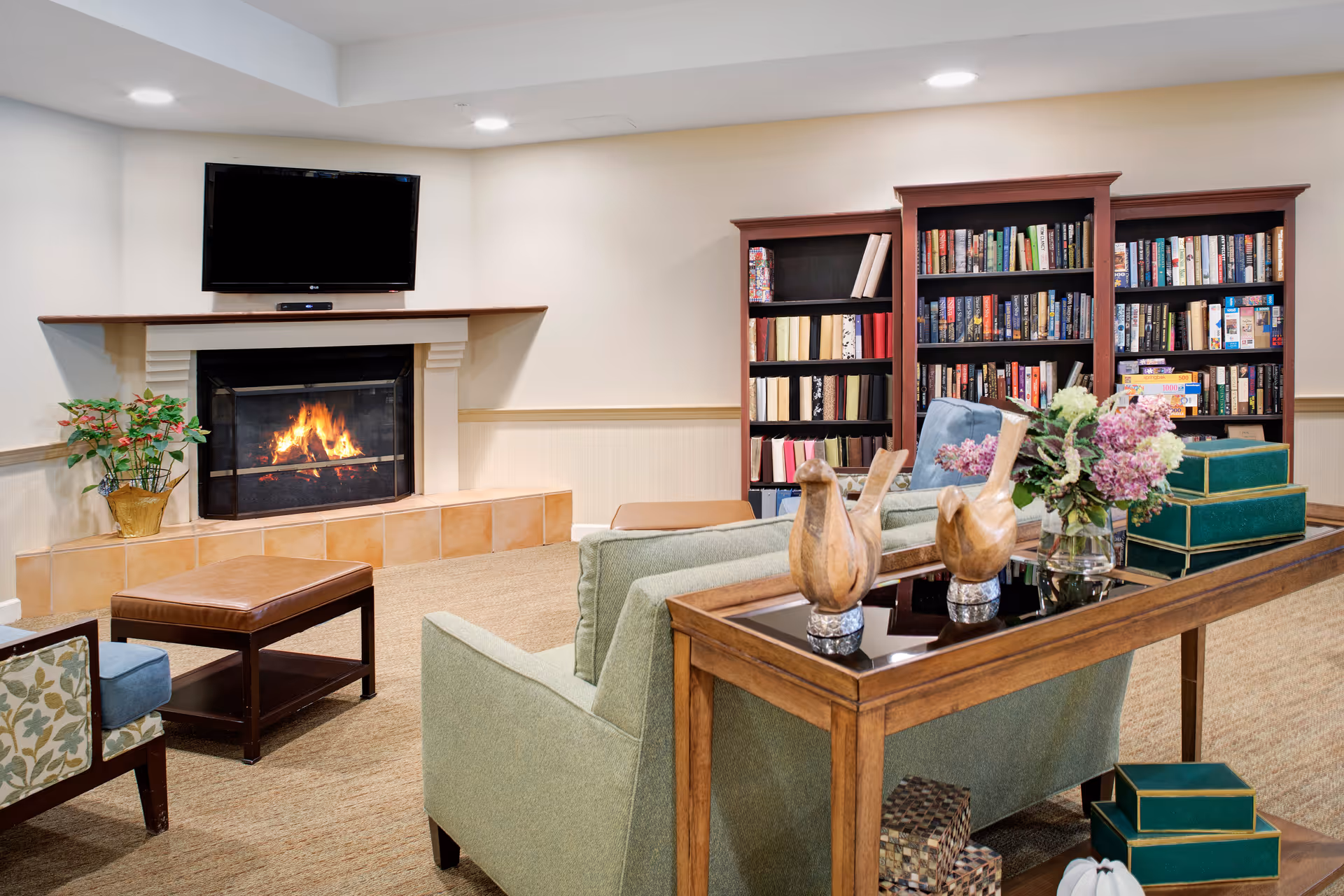 Cozy living room area with a lit fireplace beneath a wall-mounted TV, a green sofa, a wooden table with decorative wooden bird figurines and a vase of flowers, a brown ottoman, a floral-patterned armchair, and two large bookshelves filled with books.