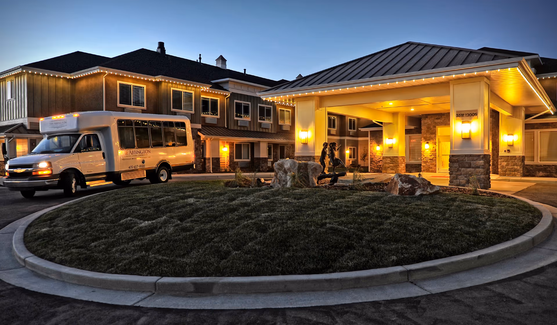 Front entrance of a senior living facility at dusk with a covered porte-cochere, a shuttle van parked nearby, and a circular driveway.