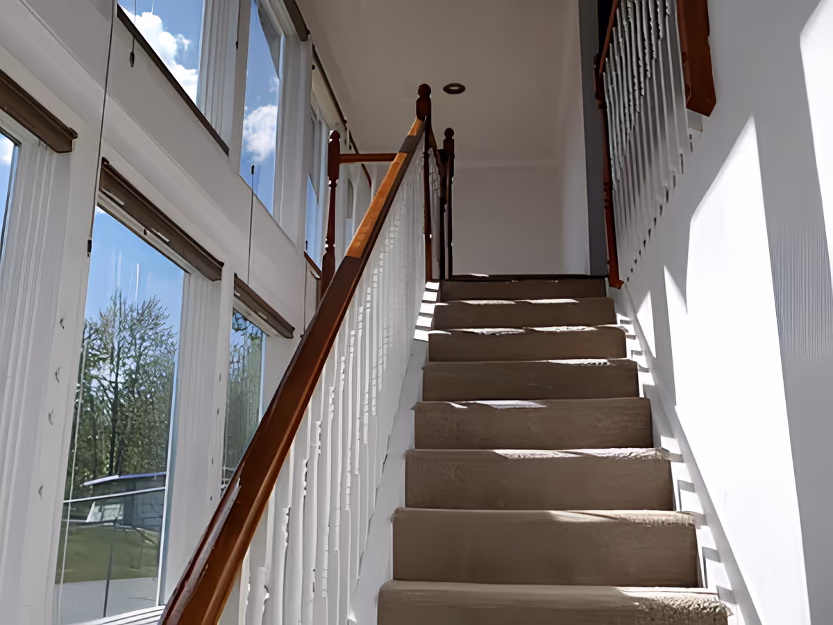 Carpeted staircase with wooden handrails on both sides, leading upwards in a bright interior space with large windows on the left side letting in natural light and showing a view of trees and a blue sky with clouds outside.