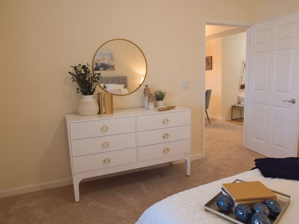 A bedroom interior featuring a white six-drawer dresser topped with plants, books and a round mirror, an open door to an adjacent room, and the corner of a bed with a decorative tray.