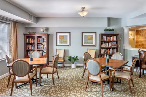 A well-lit common area with multiple wooden tables and cushioned chairs arranged on a patterned carpet. Two bookshelves filled with books flank the back wall, which also features two framed abstract artworks and a potted plant on the floor. A window with beige curtains is on the left side, and a small counter area with bar stools is visible on the right.