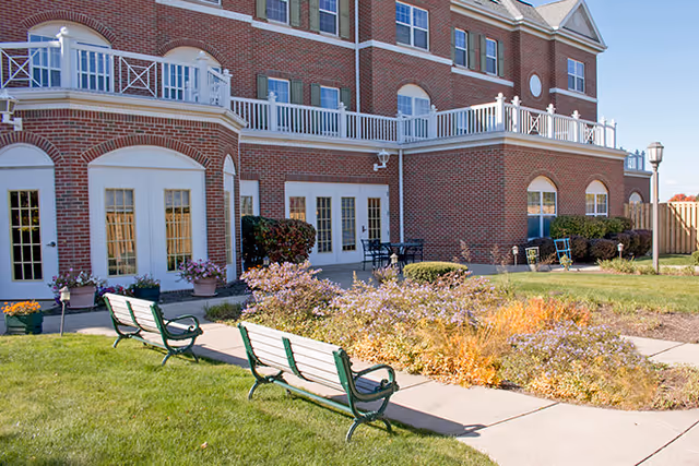 Outdoor area of a senior living facility with green benches along a concrete pathway, flower beds with purple and yellow flowers, and a brick building with white railings and multiple windows in the background.