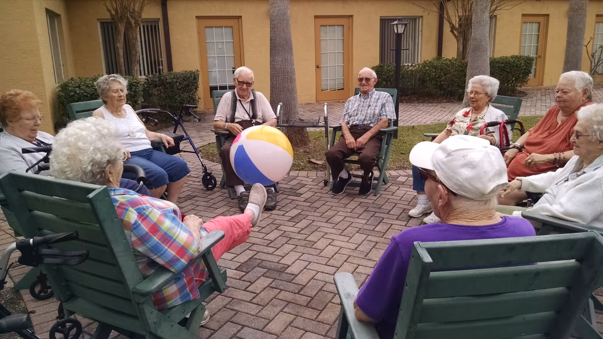 A group of elderly people sitting in a circle on green wooden chairs outdoors, playing with a colorful beach ball. The setting is a paved courtyard with yellow building walls, windows, doors, and some greenery in the background.