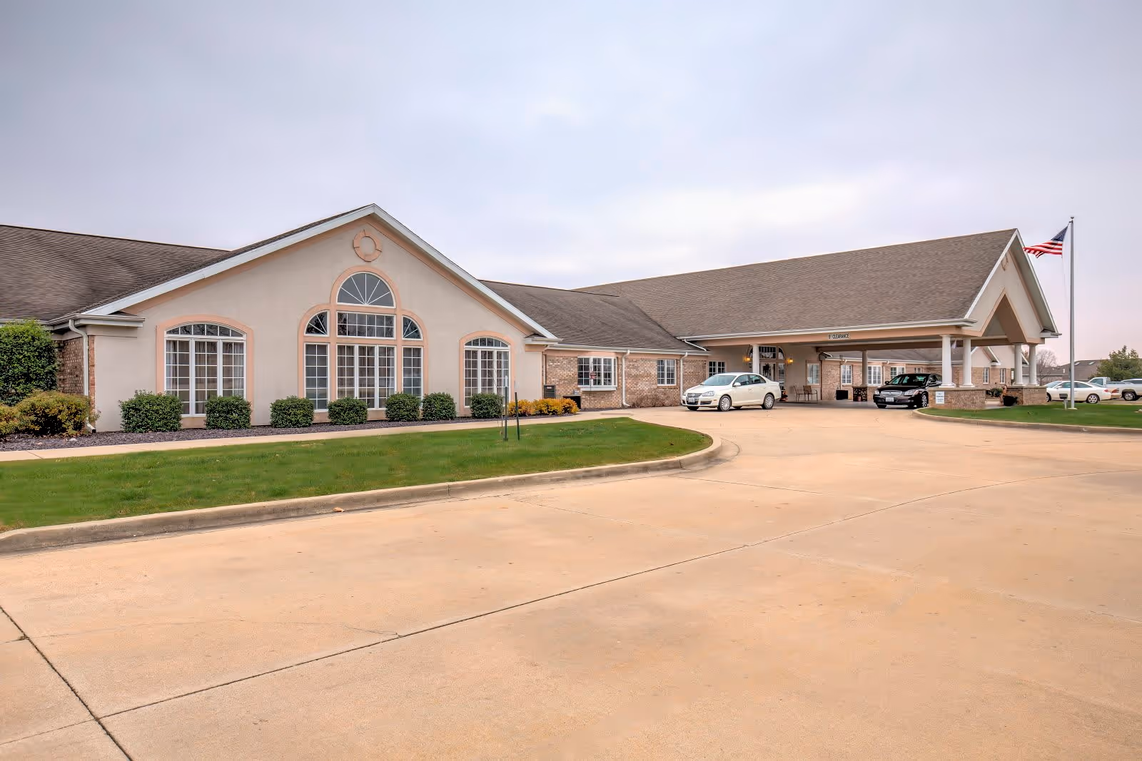 Exterior view of a single-story senior living facility building with beige walls, large windows, and a covered entrance. There are two cars parked near the entrance and an American flag on a flagpole to the right. The driveway and well-maintained lawn are visible in the foreground.