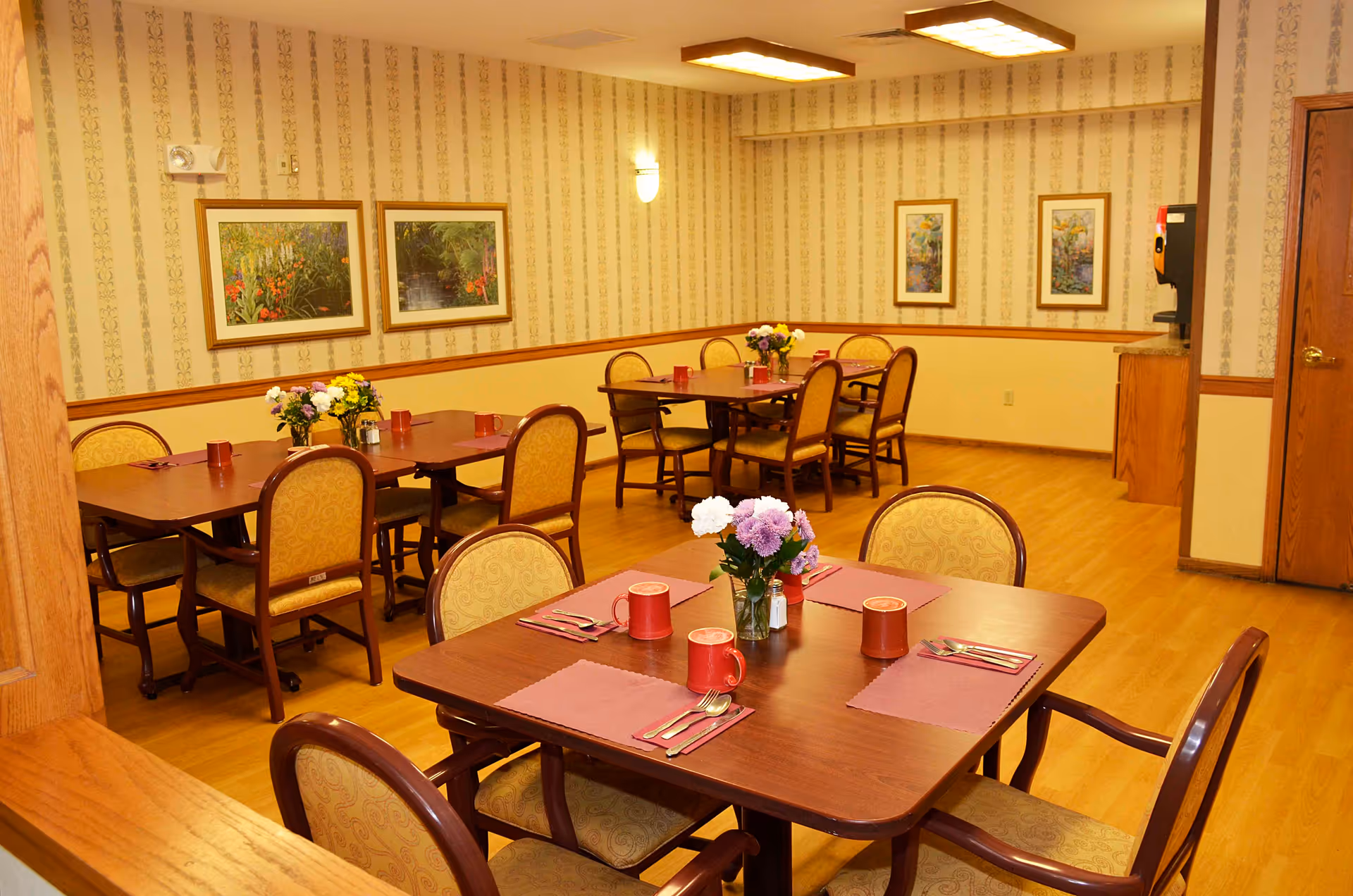 A dining room in a senior living facility with wooden tables and cushioned chairs arranged neatly. Each table is set with placemats, red mugs, and silverware. There are flower arrangements in glass vases on the tables. The walls have patterned wallpaper with framed floral artwork. The floor is wooden, and there is a beverage dispenser in the corner near a wooden door.