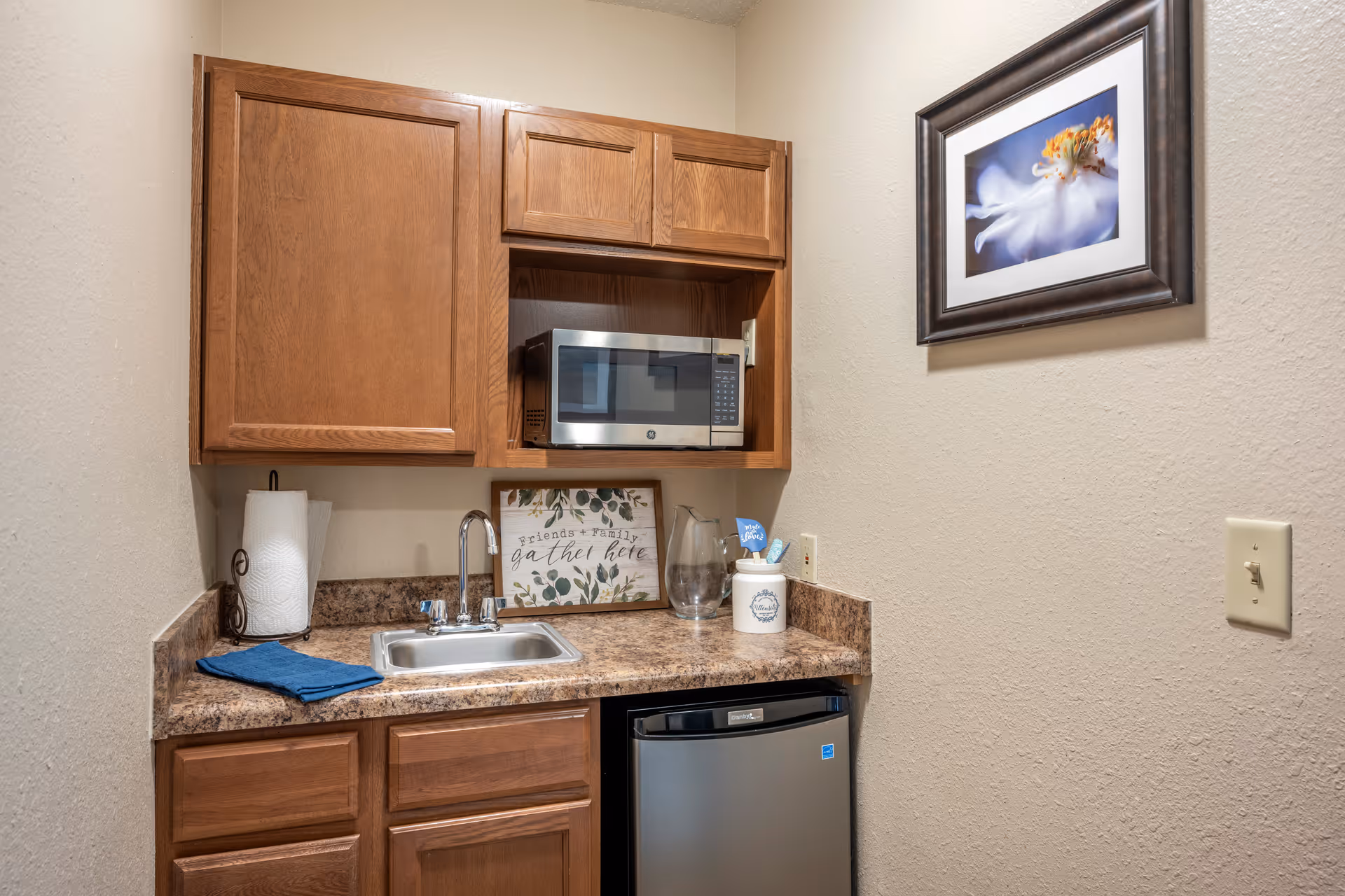 Small kitchenette with wooden cabinets, a microwave, sink, mini fridge and framed wall art.
