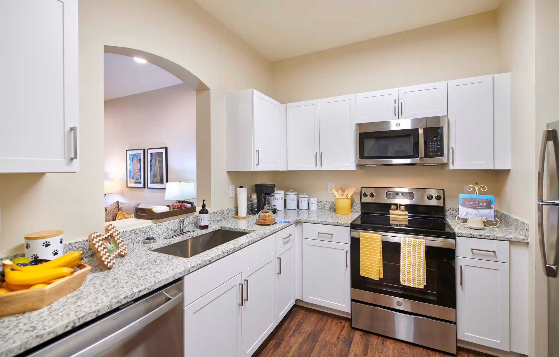 Modern kitchen with white cabinets, granite countertops, stainless steel appliances including a microwave and stove, and a sink. The kitchen has wooden flooring and is decorated with yellow kitchen towels, a coffee maker, and various kitchen accessories. An arched opening leads to a living area with framed pictures and a lamp.