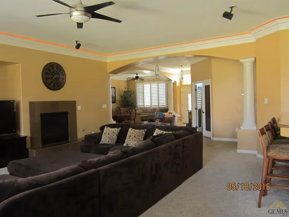 Spacious living room with a large dark sectional sofa adorned with patterned pillows, a fireplace with a large clock above it, ceiling fans, and an open archway leading to a dining area with a table and chairs. The walls are painted beige, and there is carpet flooring throughout.