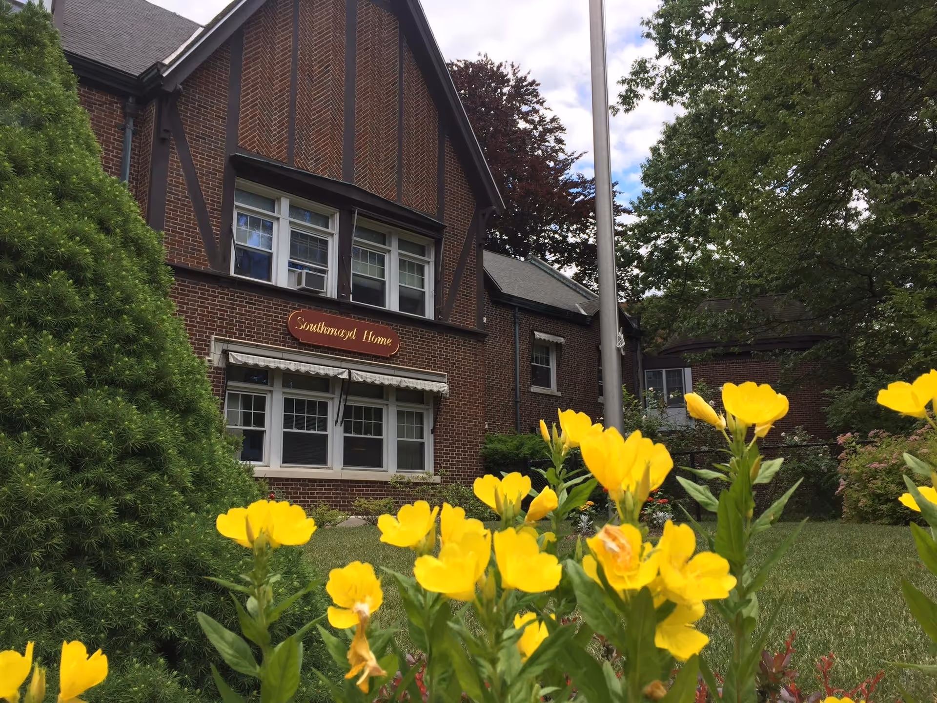 Exterior view of a brick building with a sign reading 'Southmayd Home'. The building has white-framed windows and a peaked roof. In the foreground, there are bright yellow flowers and green bushes, with trees and a flagpole nearby under a partly cloudy sky.