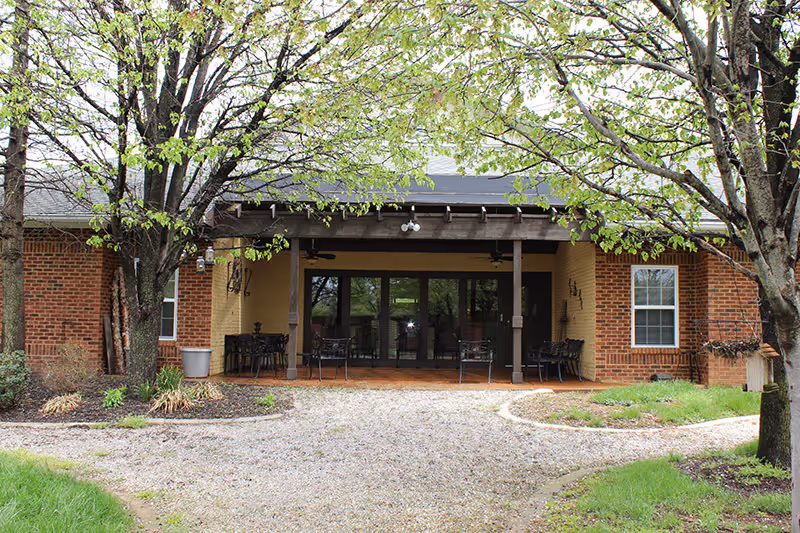 Exterior view of a single-story brick building with a covered patio area. The patio has several metal chairs and tables, and the building is surrounded by trees with fresh green leaves and landscaped garden beds. A gravel pathway leads up to the patio entrance.