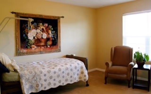 Sunny bedroom with a single bed covered by a patterned quilt, an upholstered armchair by a window, a side table with a plant, and a large floral painting on the wall.