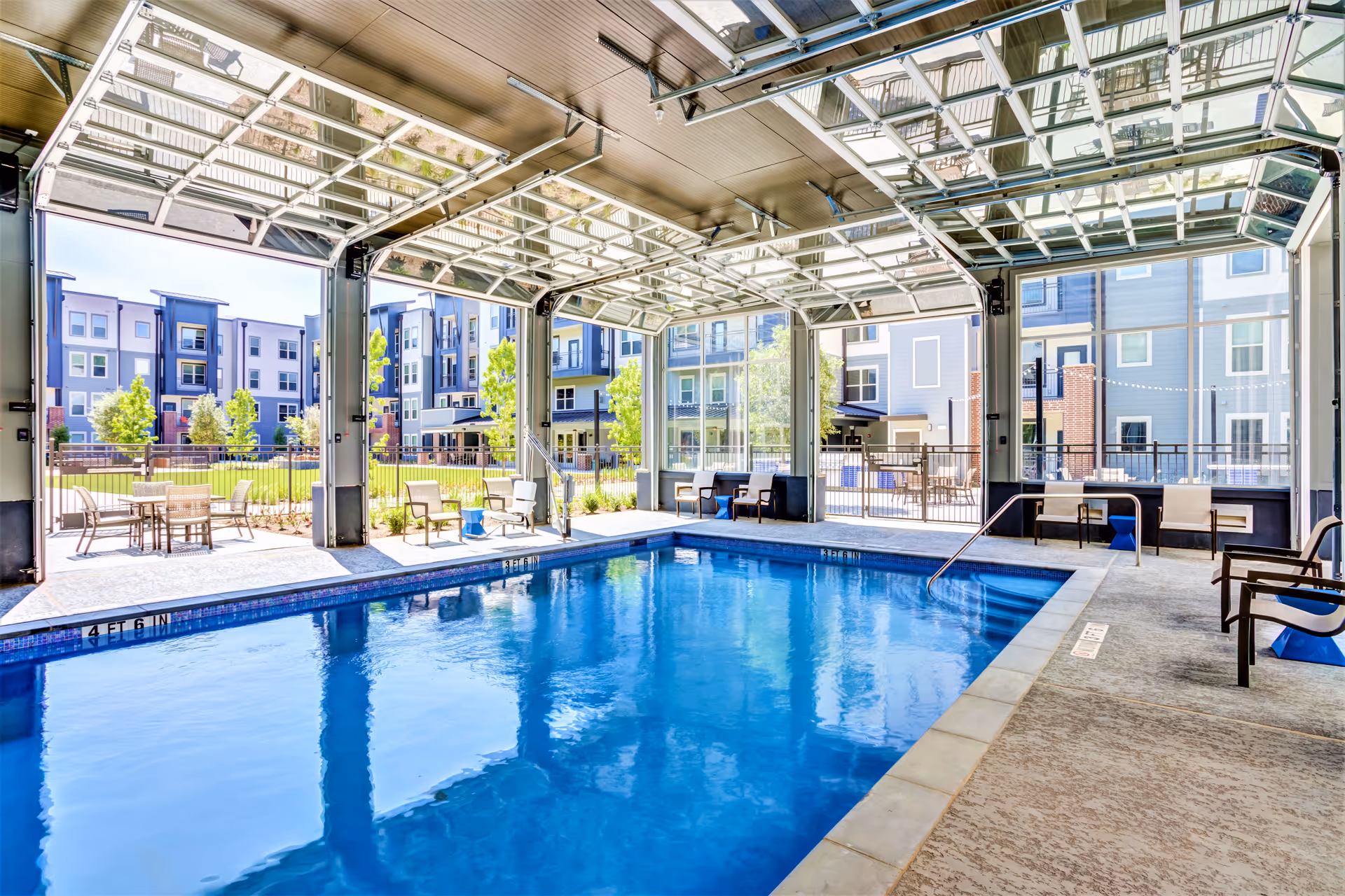 Indoor swimming pool area with large glass garage-style doors open to an outdoor patio. Several chairs and tables are arranged around the pool, and modern apartment buildings with balconies and greenery are visible outside.