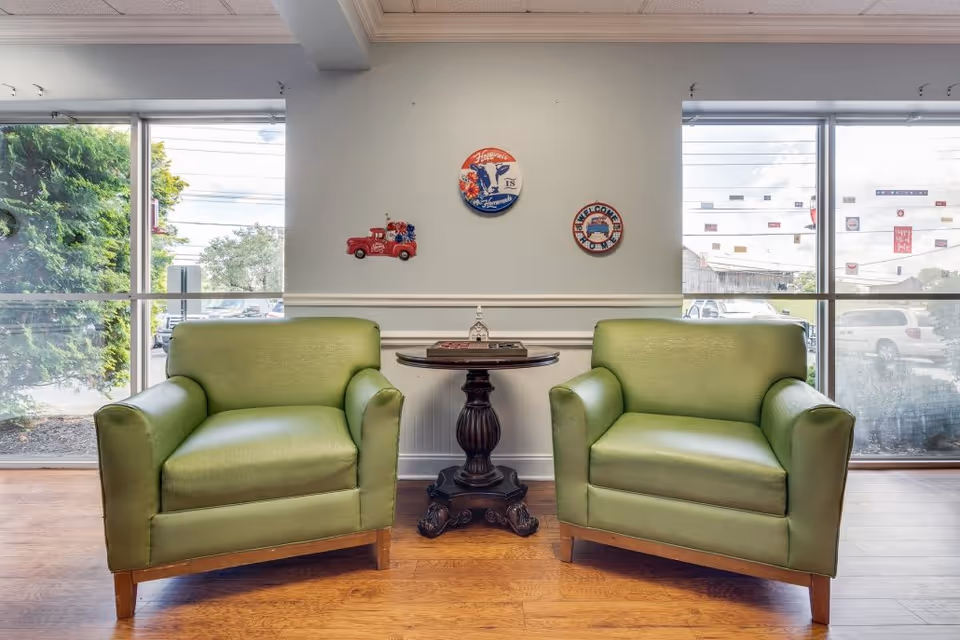 Two green armchairs with wooden legs are positioned on either side of a small round dark wooden table in a room with large windows showing an outdoor view. The wall behind the chairs has three decorative wall hangings, including a red toy truck and two circular signs with vintage designs. The floor is wooden, and the room is well-lit with natural light.