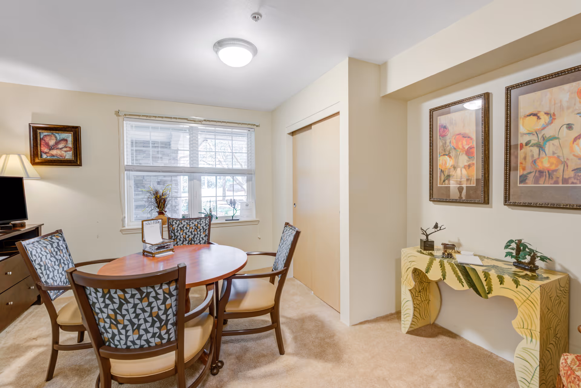 Small dining area with a round wooden table and four upholstered chairs near a window and a decorative console table with framed artwork.