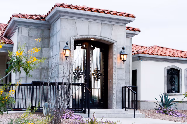 Front exterior view of a building with a tiled roof, stone facade, and decorative wrought iron double doors. There are two wall-mounted lantern-style lights on either side of the entrance, with plants and landscaping in the foreground.