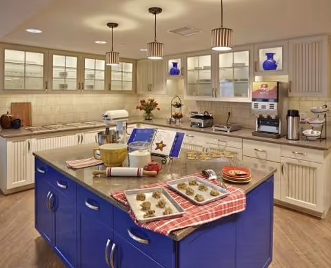 A bright and clean kitchen area with white cabinets and glass doors, a blue island in the center with baking trays holding cookies, a rolling pin, a mixing bowl, and a cookbook stand. The countertop has various kitchen appliances including a coffee maker and a beverage dispenser. Pendant lights hang from the ceiling, and decorative blue vases are displayed in the cabinets.