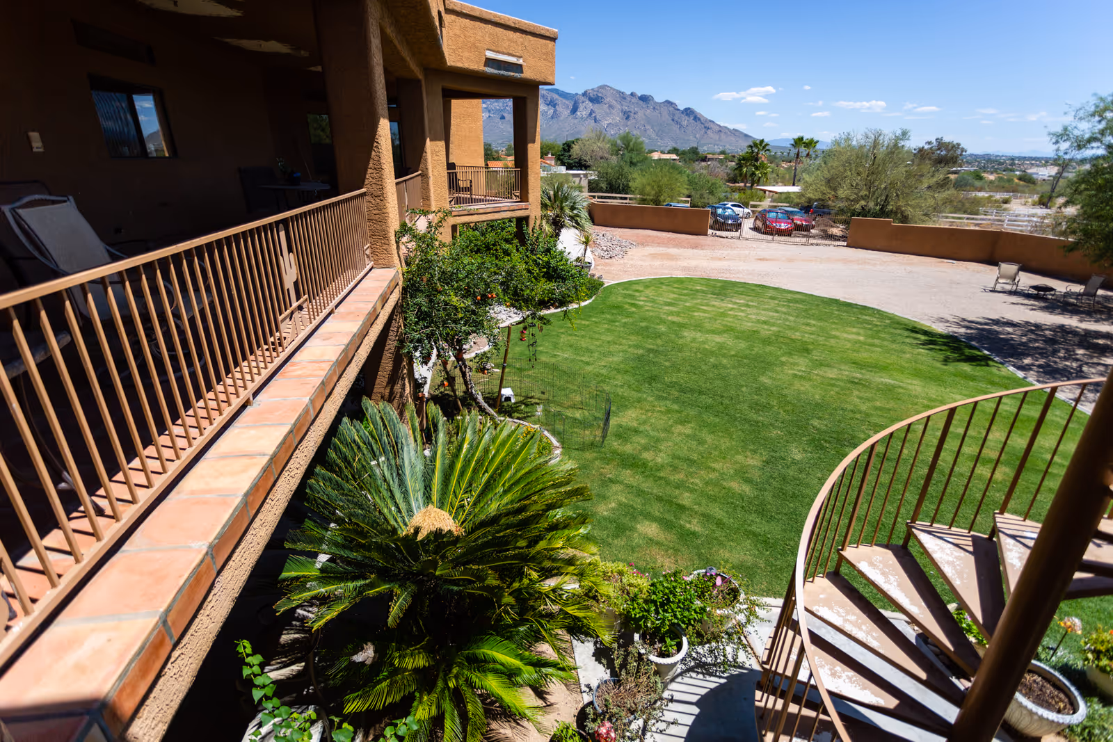 View of a green lawn area surrounded by a brown building with a balcony and a spiral staircase. There are plants and trees around the lawn, with mountains visible in the background under a clear blue sky.