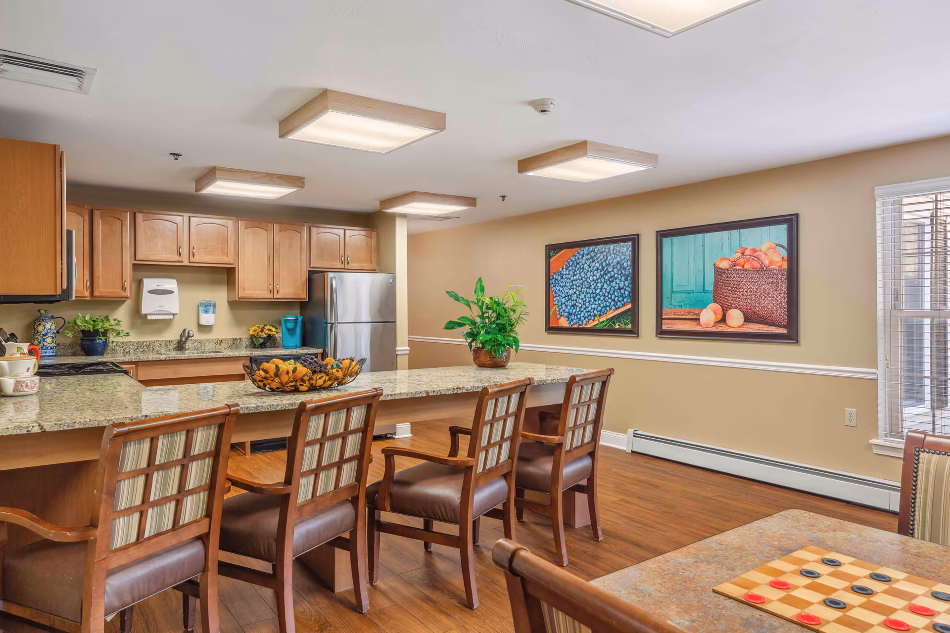 A bright and spacious kitchen area in a senior living facility featuring wooden cabinets, a granite countertop island with four wooden chairs, a stainless steel refrigerator, and decorative plants. Two framed pictures of fruit hang on the beige wall, and a window with blinds allows natural light to enter. In the foreground, a table with a checkers game is partially visible.
