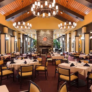 A spacious dining room with round tables covered in white tablecloths, each set with plates, glasses, and black napkins. The room features a high wooden ceiling with exposed beams and multiple large chandeliers. A stone fireplace is centered on the far wall, flanked by tall windows and plants, creating a warm and inviting atmosphere.