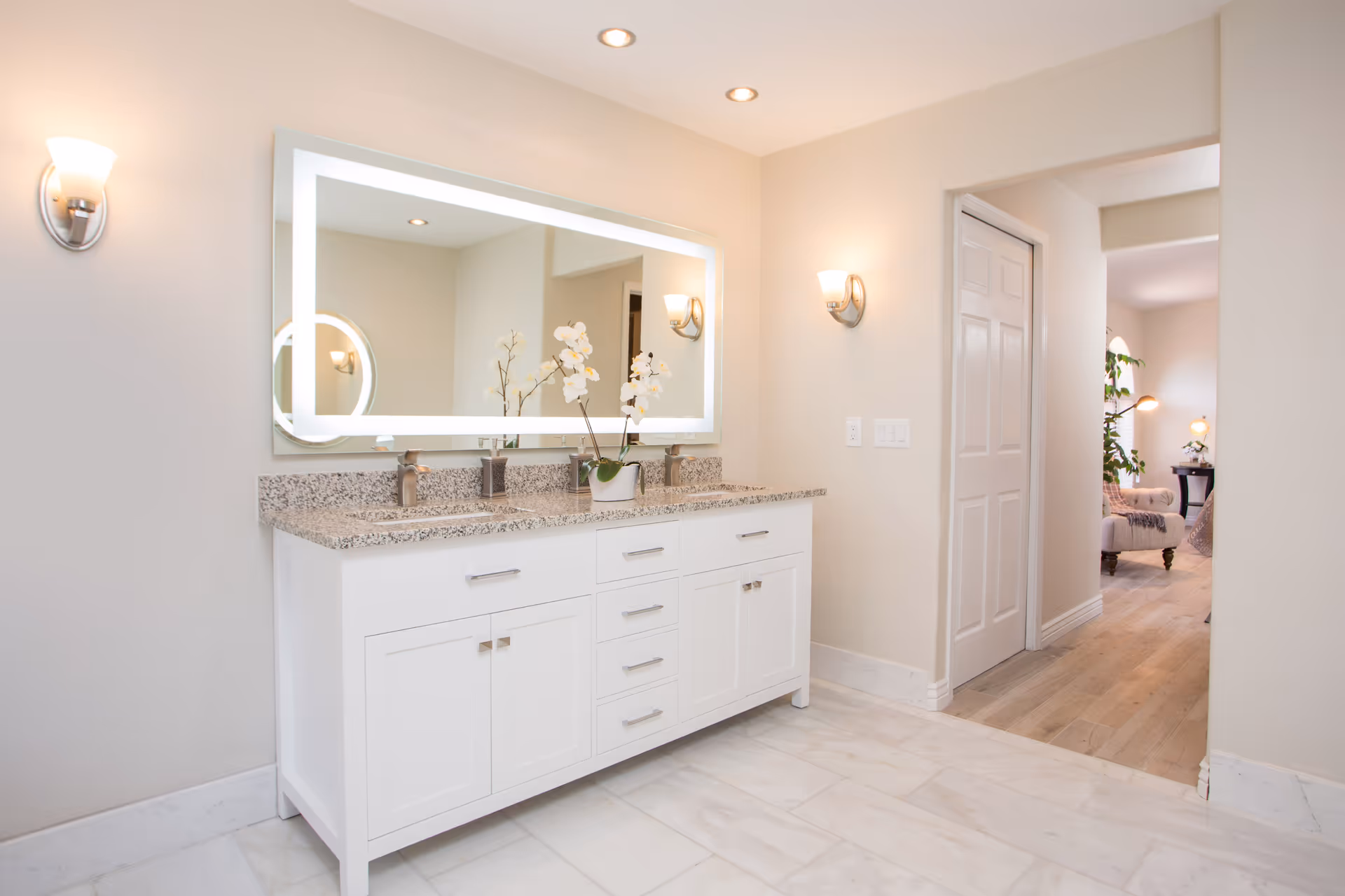 A bright and clean bathroom featuring a large illuminated rectangular mirror above a white double sink vanity with granite countertop. The vanity has multiple drawers and cabinets. Two wall-mounted light fixtures flank the mirror. The floor is tiled in white marble, and an open doorway leads to a living area with wooden flooring and furniture visible in the background.