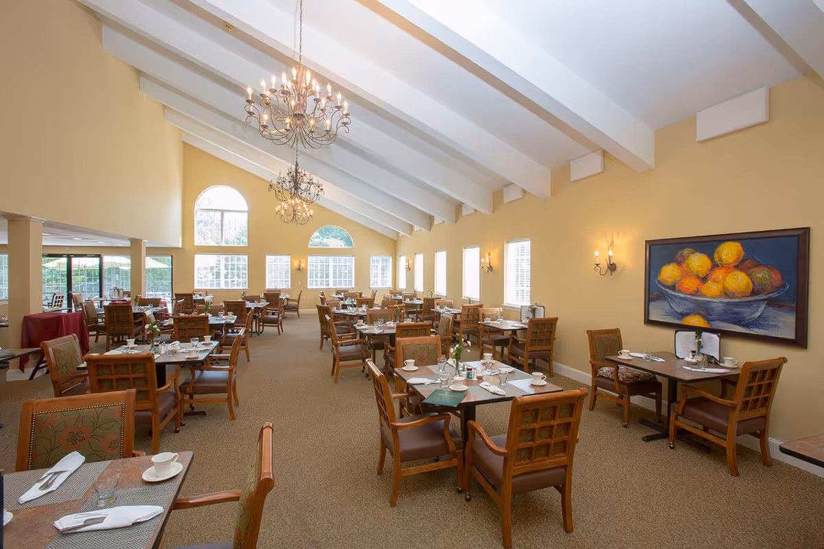 Large, bright dining room with multiple set tables and chairs, chandeliers, high beamed ceiling, and a fruit painting on the wall.