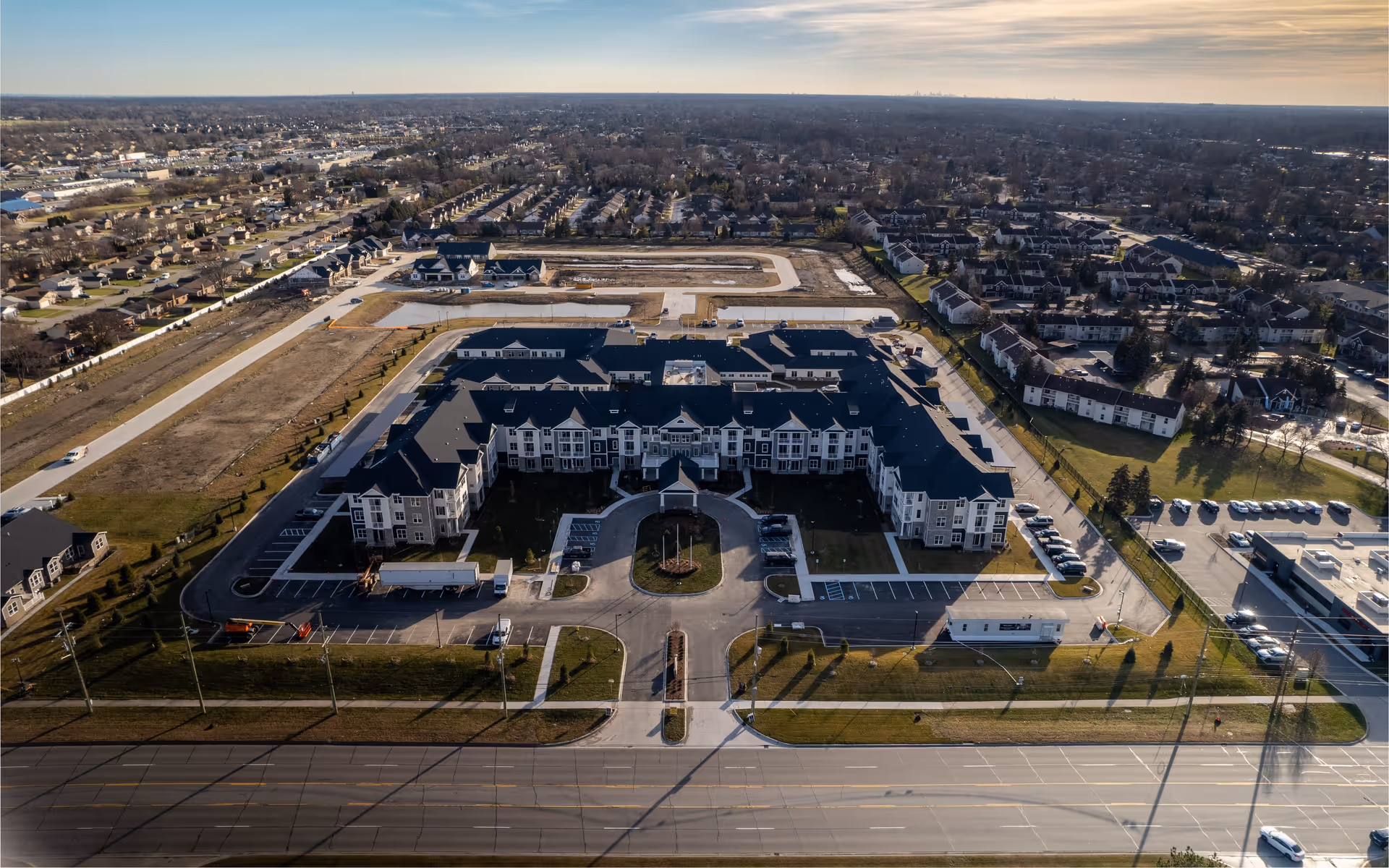 Aerial view of a large senior living facility building with a U-shaped layout, surrounded by parking lots and roads. The facility is situated in a suburban area with many houses and other buildings visible in the background under a partly cloudy sky.
