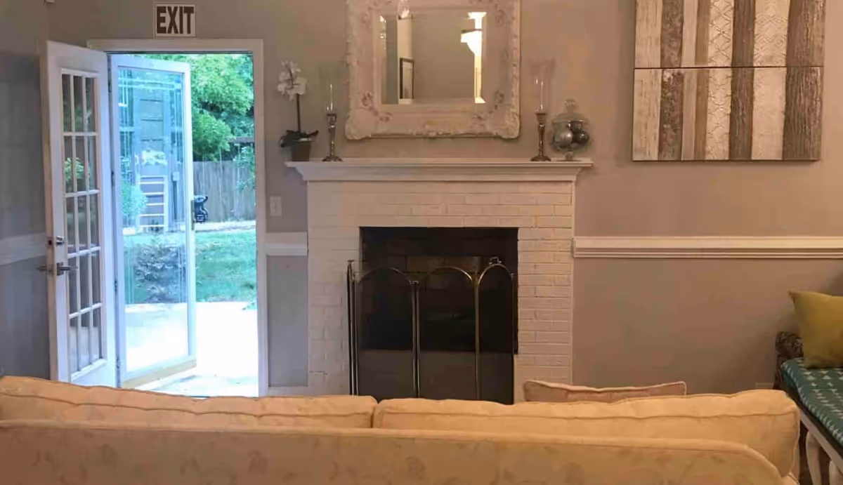 Interior view of a senior living facility common area with a beige couch in the foreground, a white brick fireplace with a decorative mirror and candlesticks on the mantel, a piece of abstract wall art, and an open door leading outside to a green yard.