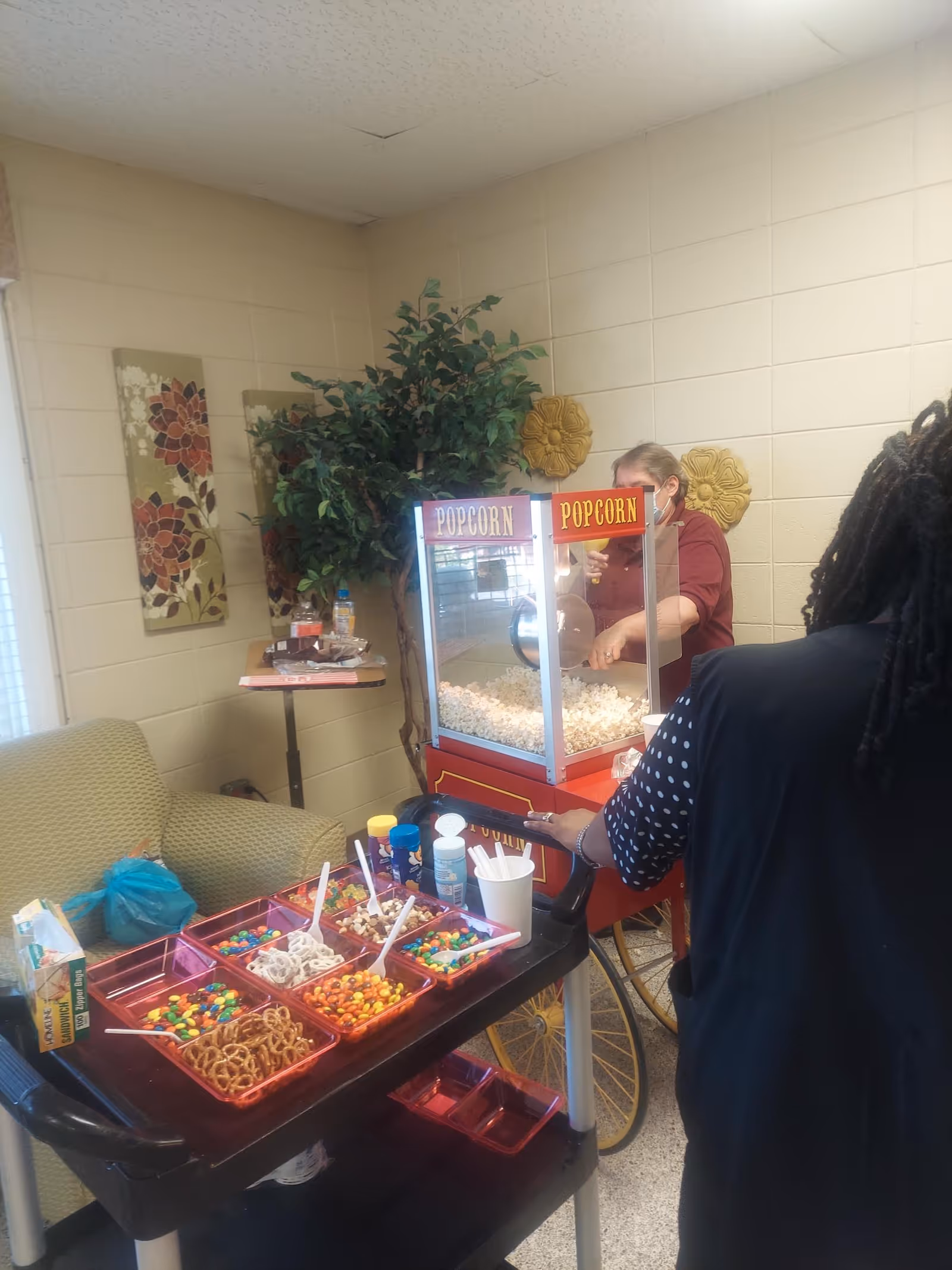 A popcorn machine and cart with a tray of candy toppings in a senior living common room while staff serve residents.