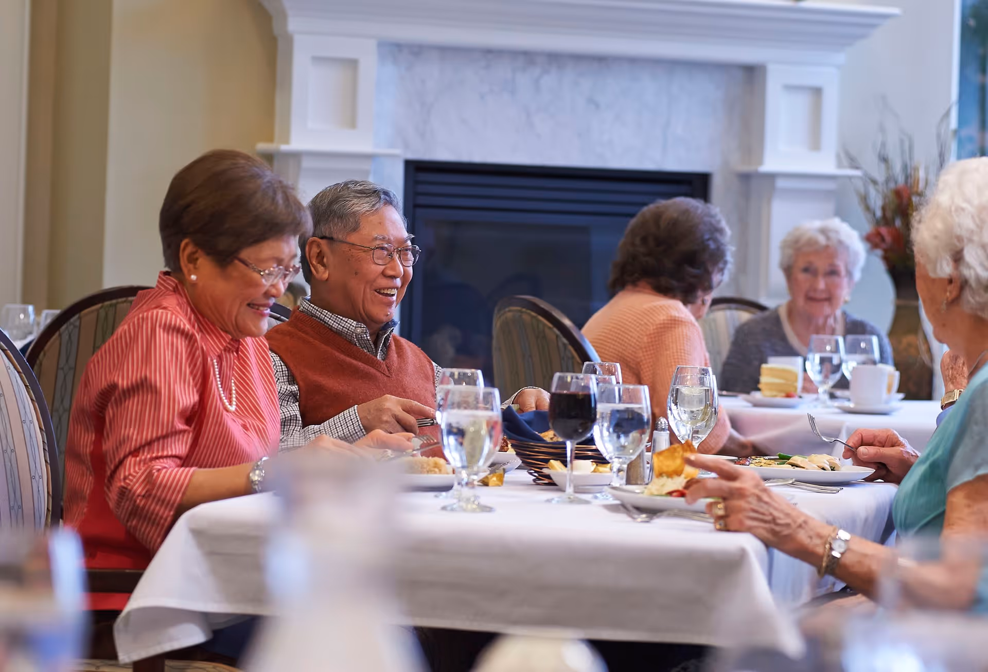 A group of elderly people sitting around a dining table in a well-lit room with a fireplace in the background, enjoying a meal and engaging in conversation.