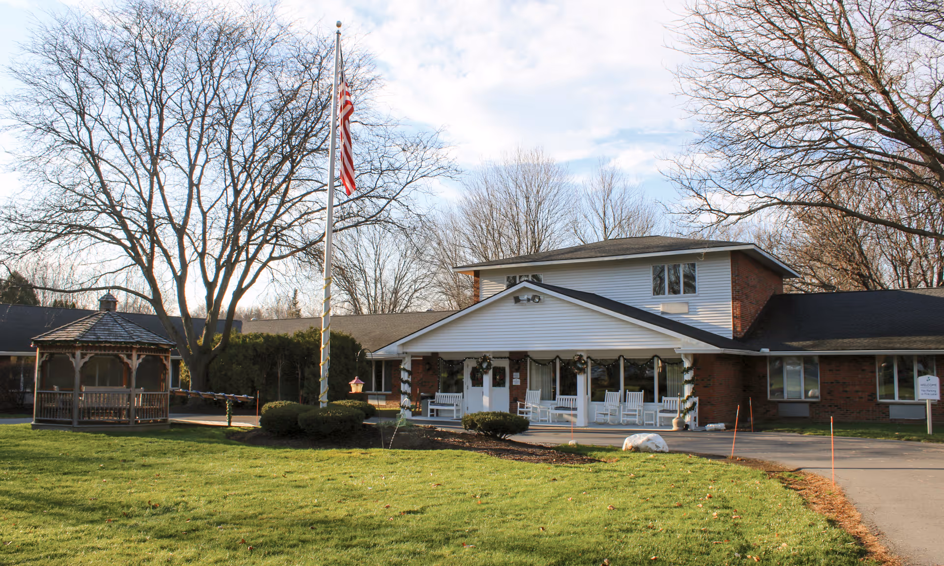 Exterior view of a senior living facility building with a covered porch featuring white rocking chairs. There is a flagpole with an American flag in front, a small gazebo to the left, and leafless trees surrounding the area. The grass is green and the sky is partly cloudy.
