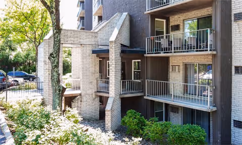 Exterior view of The Guardian Apartments showing a multi-story building with balconies, surrounded by trees and shrubs, with a parking area visible in the background.