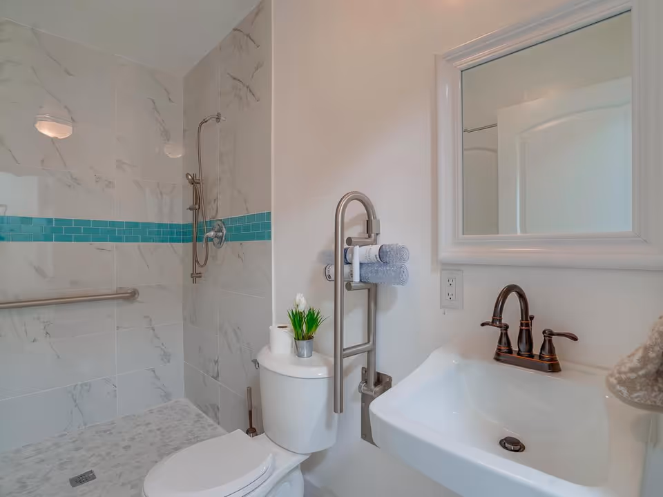 A clean accessible bathroom featuring a walk-in tiled shower with a turquoise tile accent, a toilet with a grab bar and towel rack, and a sink with mirror.