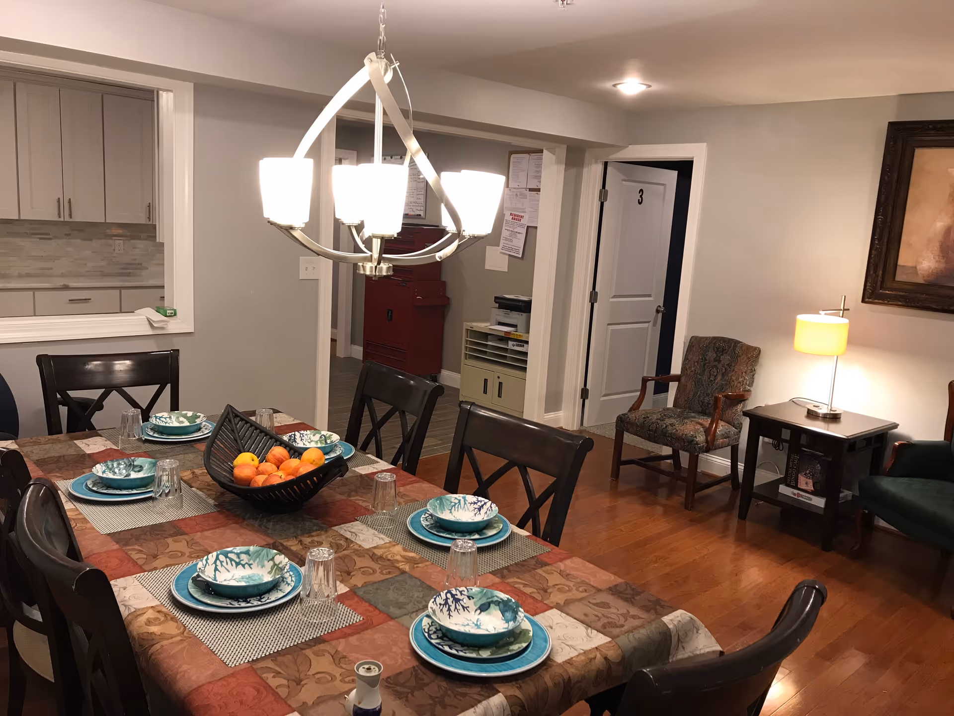 Dining room with a table set for six people with plates, bowls, and glasses. A decorative bowl with fruit is in the center of the table. There is a modern chandelier hanging above the table. In the background, there is a doorway leading to a hallway with a red cabinet and a white door labeled with the number 3. To the right, there are two chairs, a side table with a lamp, and a framed painting on the wall. The floor is wooden, and the walls are painted light gray.
