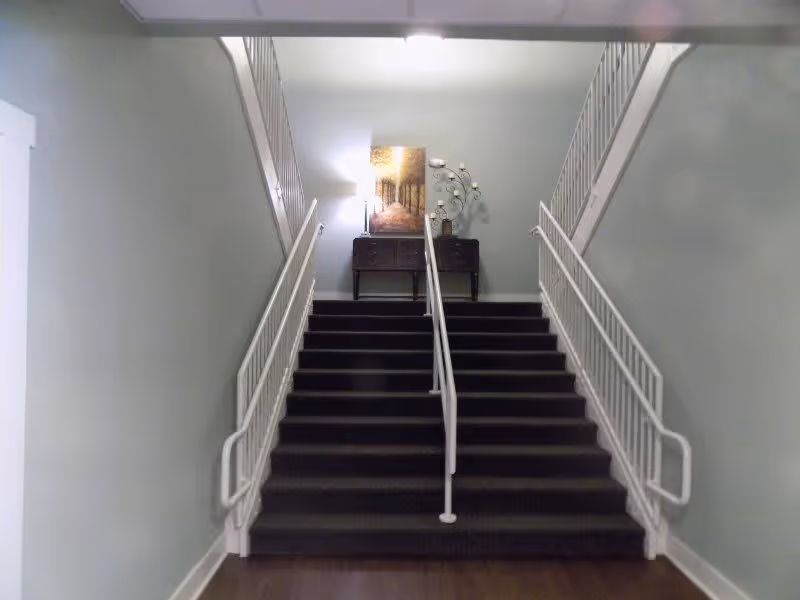 Indoor staircase with dark carpeted steps and white railings on both sides and in the middle. At the top of the stairs, there is a small dark wooden table with a lamp, a decorative tree-like candle holder, and a framed picture of a forest path. The walls are painted light gray.