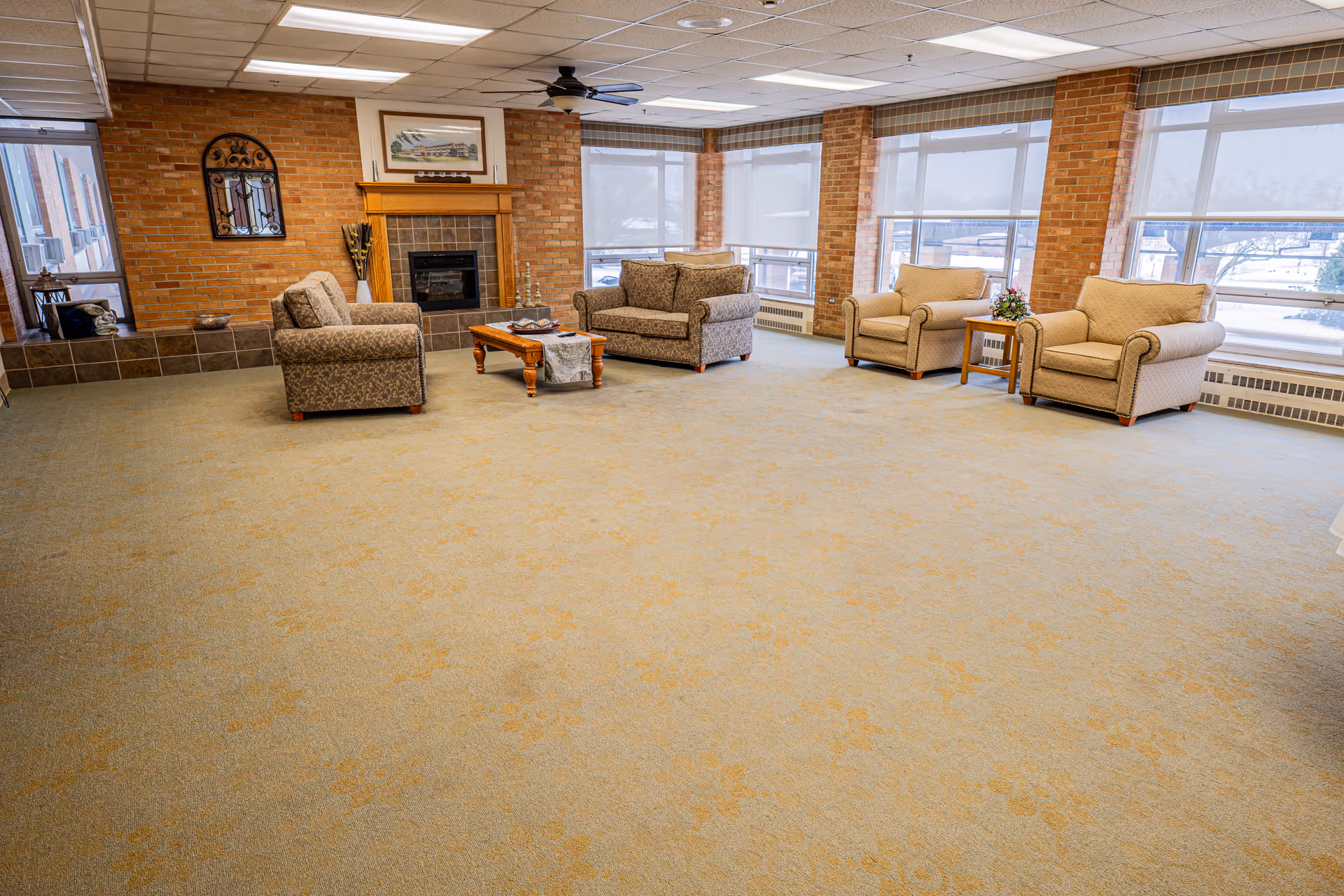 A spacious living room area with large windows letting in natural light. The room features a beige carpet with a subtle pattern, four upholstered armchairs arranged around a wooden coffee table, and a fireplace with a wooden mantel against a brick wall. There is a ceiling fan above and decorative items on the mantel and side table.