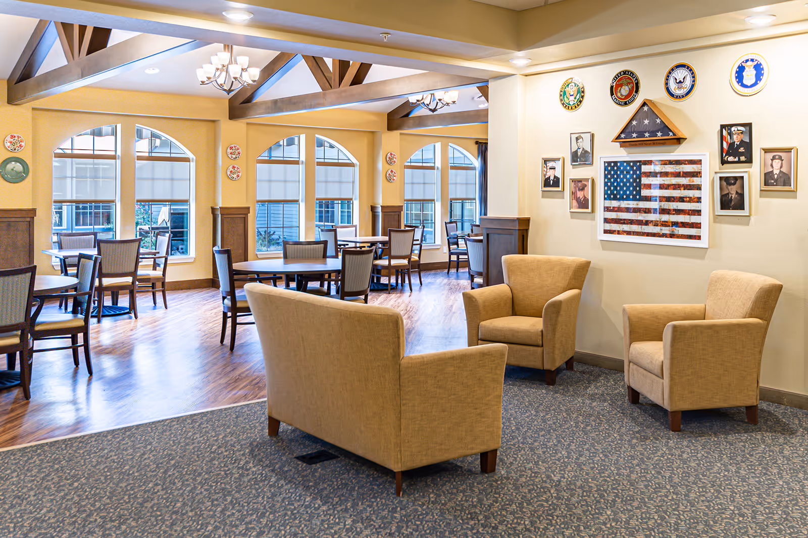 A bright and spacious common area in a senior living facility featuring beige armchairs arranged around a small seating area with carpeted flooring. In the background, there is a dining area with round tables and chairs on wooden flooring, large arched windows with blinds, and exposed wooden ceiling beams. On the wall, there is a display of military emblems, framed photographs, and a folded American flag in a triangular case.