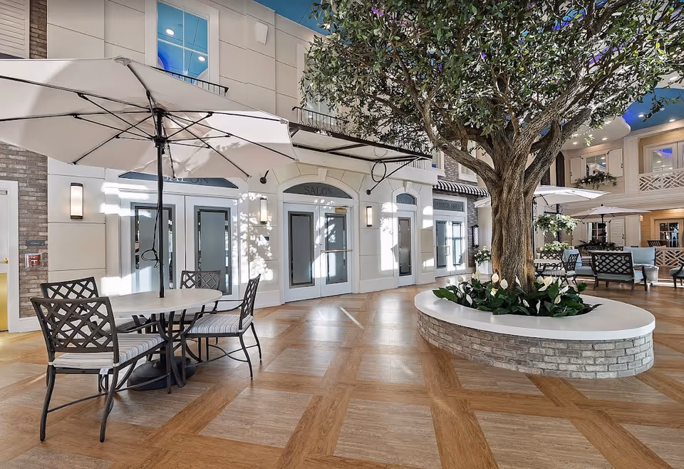 Indoor common area with a large tree planted in a circular brick planter. There are tables with umbrellas and chairs arranged around the space. The background shows doors labeled 'Salon' and 'Barber Shop' with a bright and airy atmosphere.