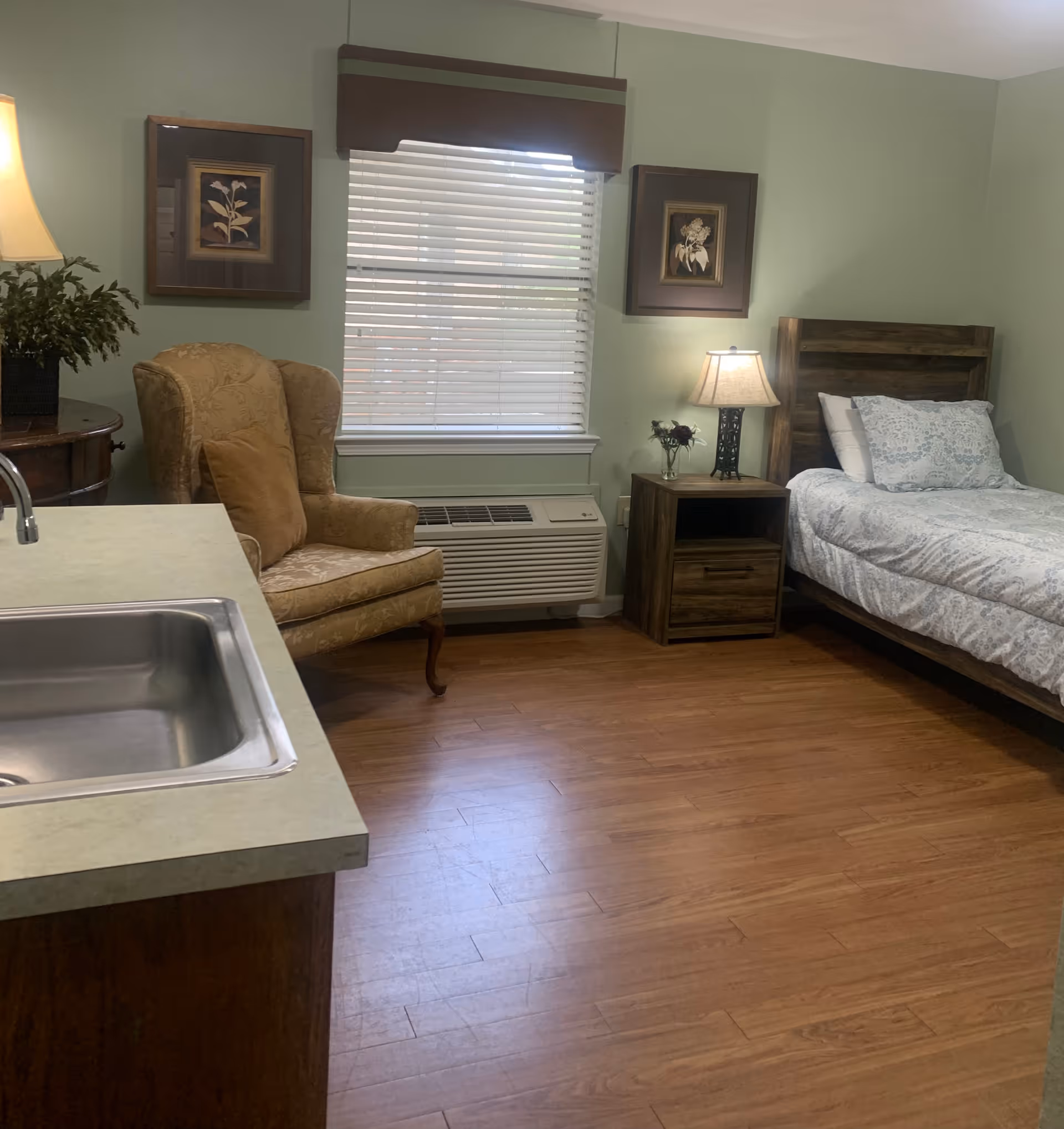 A cozy assisted living room with a single bed featuring white and blue patterned bedding, a wooden nightstand with a lamp and a small vase of flowers, a comfortable upholstered armchair with a brown cushion, a window with blinds and a brown valance, two framed floral artworks on the light green walls, and a countertop with a stainless steel sink in the foreground.