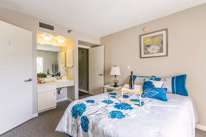 A bedroom in a senior living facility with a bed covered in a white and teal floral bedspread, a small wooden tray with a bottle and glass on the bed, a bedside table with a lamp, a framed floral artwork on the wall, an open door leading to a hallway, and a vanity sink area with a mirror and lights.